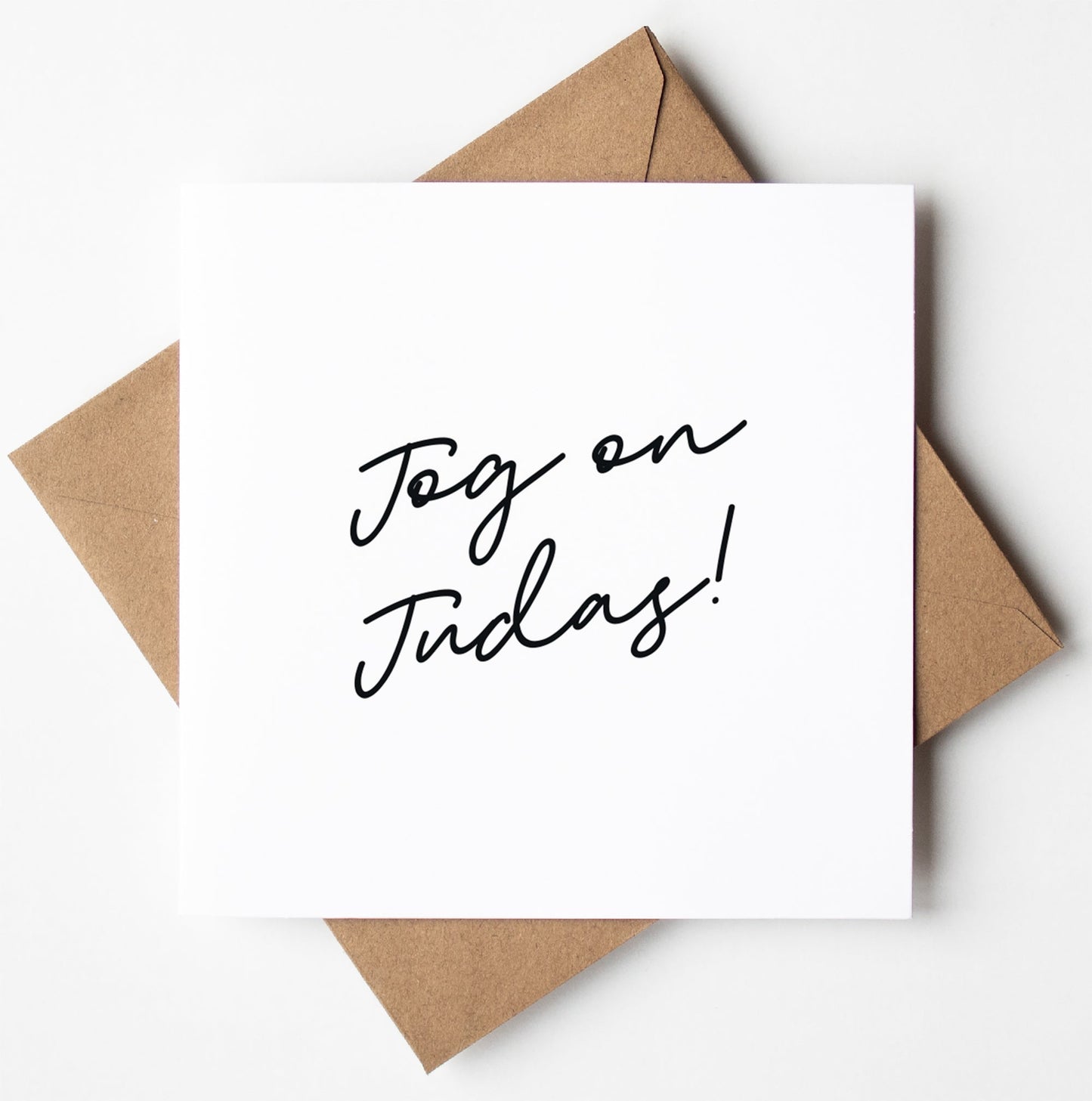 The "Jog On Judas Script Card" in white, featuring the handwritten message "Jog on Judas!", is placed on a brown envelope against a white background, making it the perfect cheeky farewell card for an unforgettable departure.