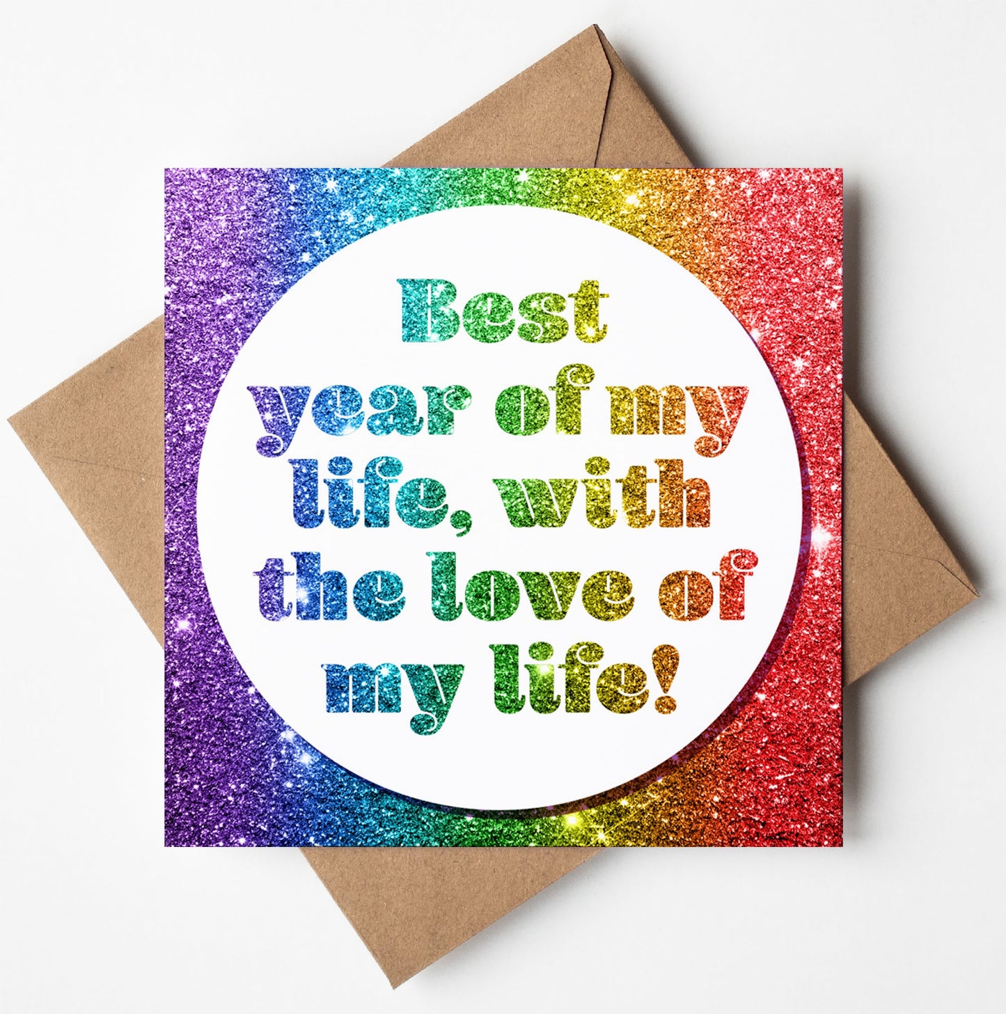 The "Best Year Of My Life Card" features a vibrant rainbow glitter background with a white circle in the center. The text on the card says, "Best year of my life, with the love of my life!" It's an ideal card for celebrating relationship goals, presented on a brown envelope and left blank inside for your personal message.