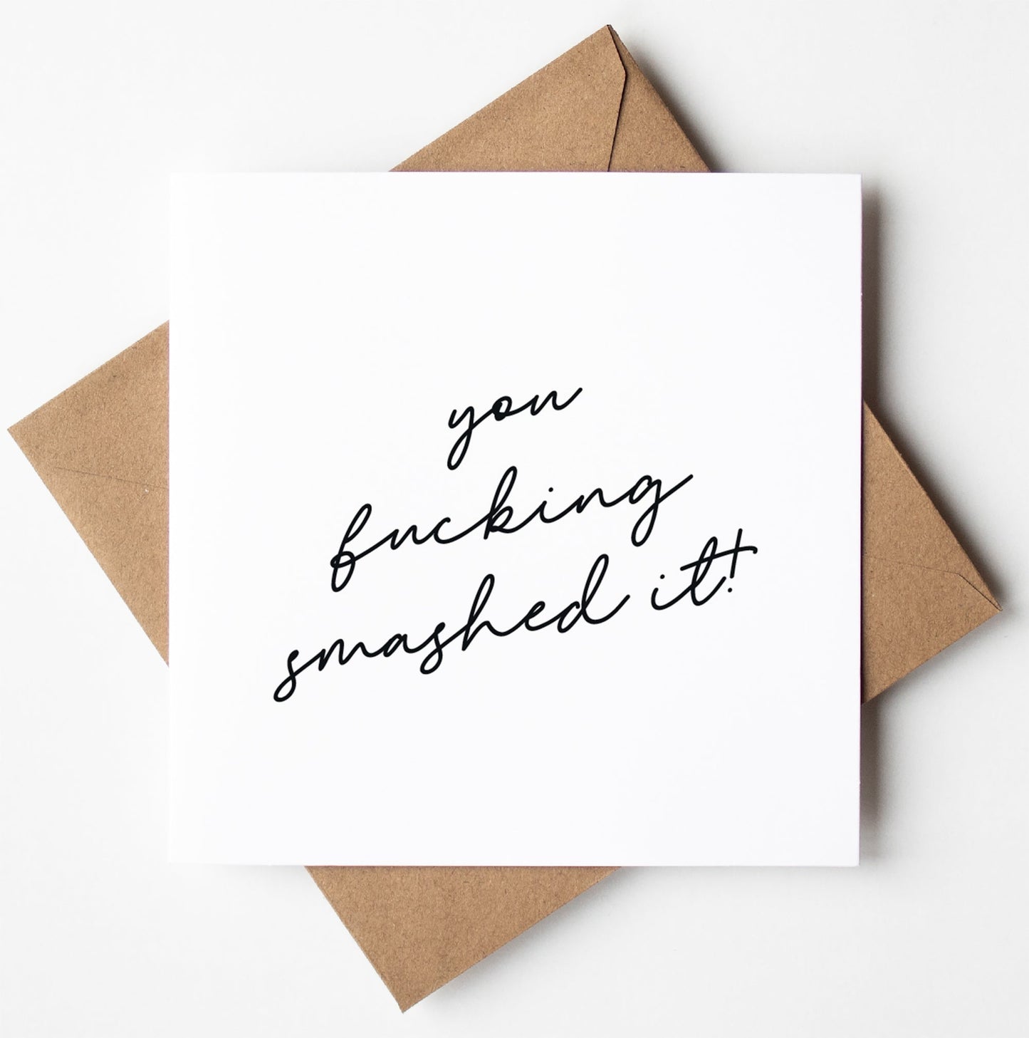 The You Fucking Smashed Script Card showcases a white greeting card with "you fucking smashed it!" elegantly written in black cursive. It is paired with a brown envelope, set against a white background, and is now available for convenient worldwide shipping.
