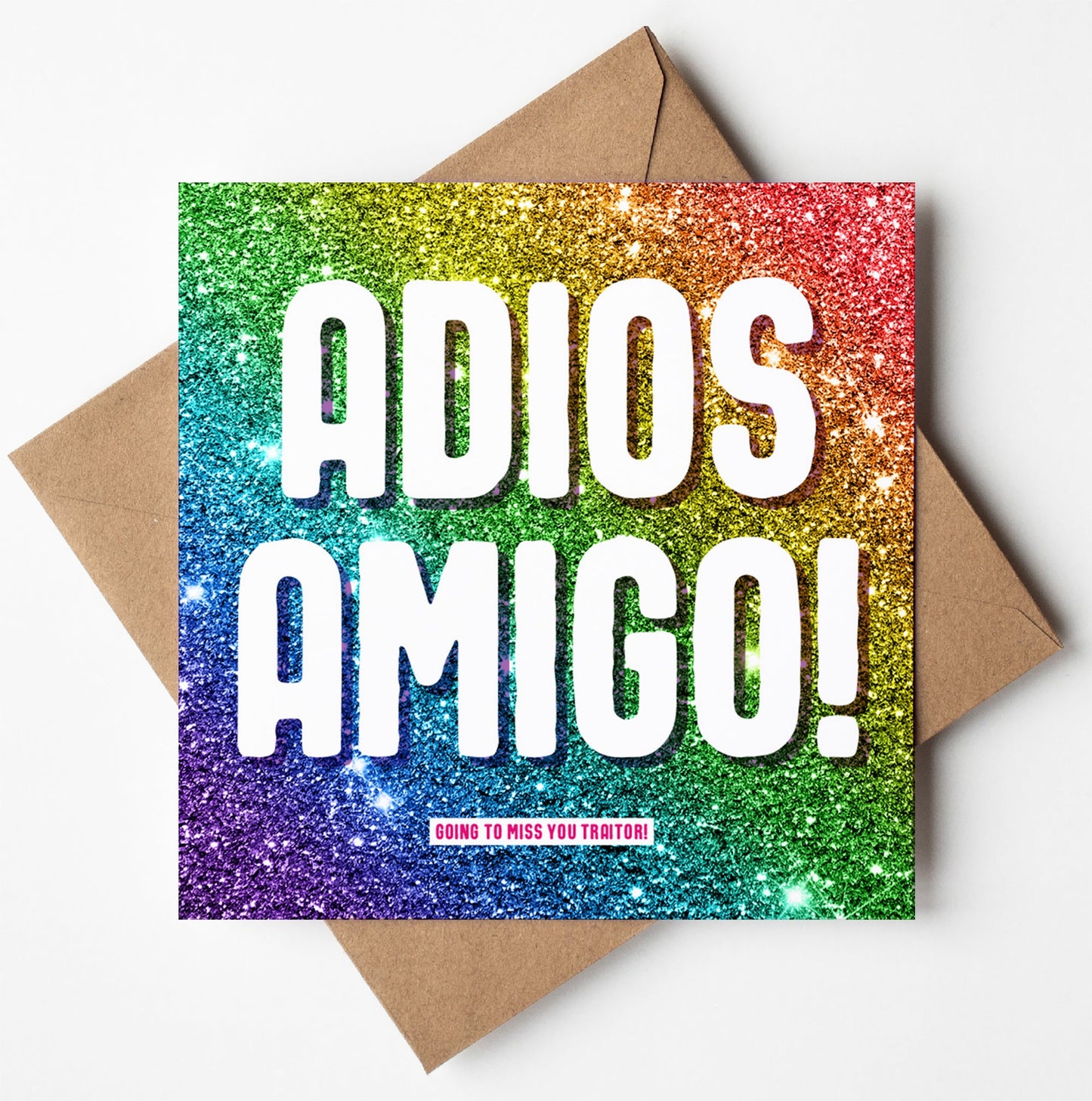 Colourful glitter greetings card with "ADIOS AMIGO!" in bold white letters and small pink text saying "GOING TO MISS YOU TRAITOR!" beneath. The artwork is playfully placed on a brown envelope.
