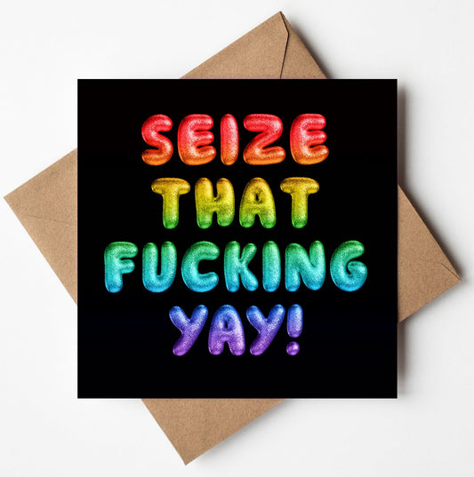 The "Seize That Fucking Yay! Rainbow Font Card" features bold, colorful bubble letters in a vibrant gradient set against a black background. This biodegradable card comes with a brown envelope and is available for worldwide shipping.