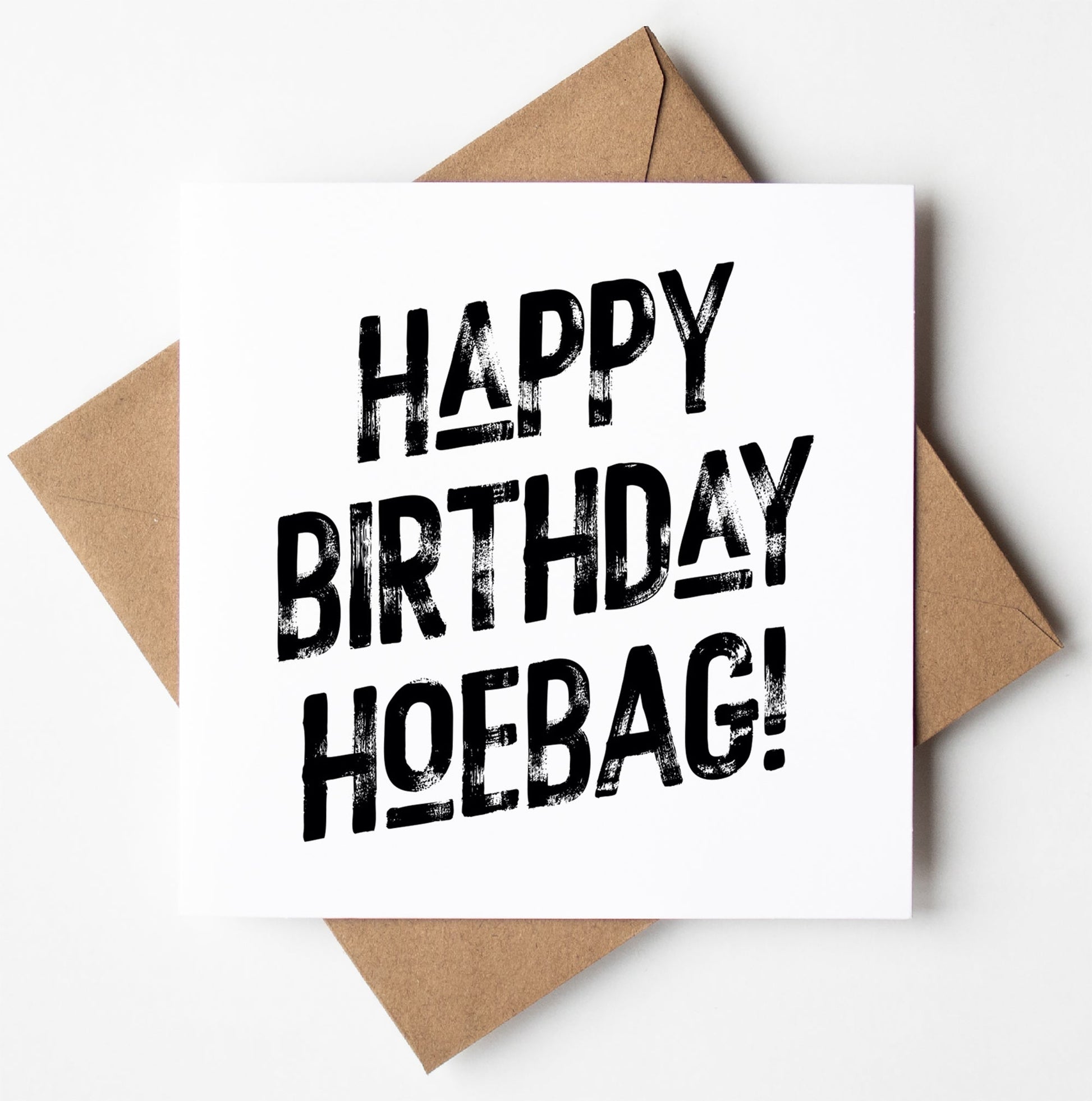 A card named "Happy Birthday Hoebag! Harlem Card" featuring humorous, bold, distressed black lettering rests on top of a brown envelope against a white background.