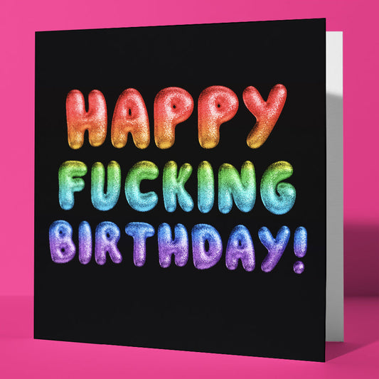 The Happy Fucking Birthday! Rainbow Font Card features bold, colorful balloon-style letters on a black background spelling out the message, making it a hilarious birthday card that's sure to bring laughs. Displayed on a pink surface.