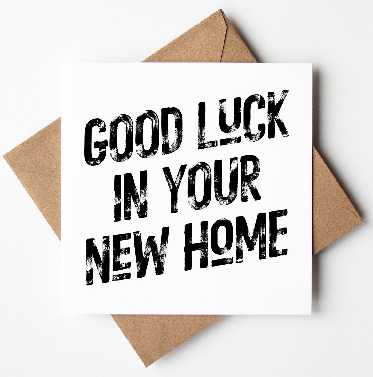 A "Good Luck in Your New Home Congratulations Card," featuring bold black text on a white background, is perched atop a brown envelope.