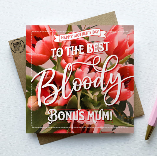 The "Happy Mother's Day To The Best Bloody Bonus Mum!" card, adorned with pink flowers and resting on a brown envelope beside a pink pen, is poised to spread love and cheer.