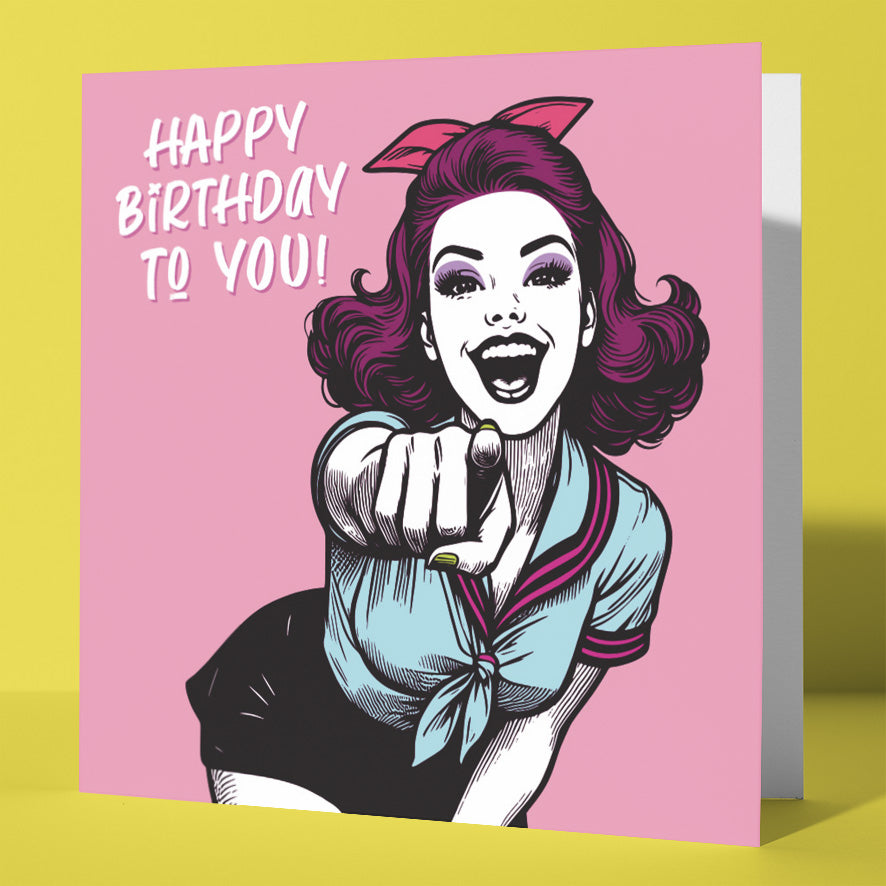 The Happy Birthday To You! Vintage Babes Birthday Card features a retro illustration of a smiling, purple-haired woman with a bow, pointing forward on a pink background with playful birthday text—perfect for any celebration.