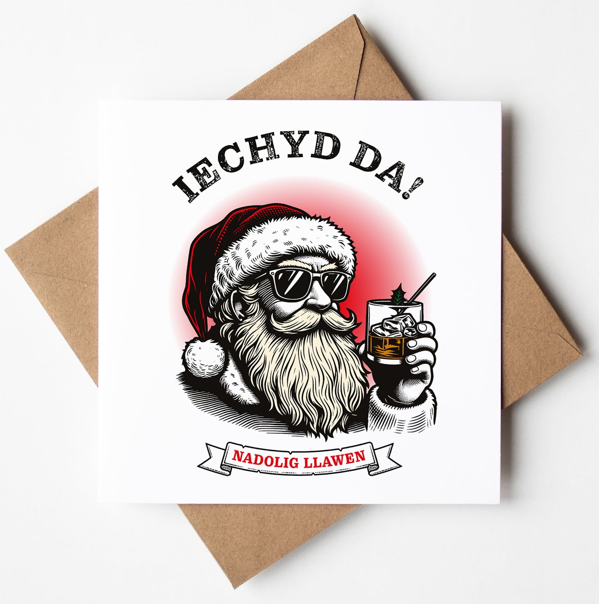 Illustration of Santa wearing sunglasses and holding a drink, with "Iechyd Da!" above and "Nadolig Llawen" below—perfect as a Welsh Christmas card. The backdrop is white with a brown envelope, ideal for worldwide shipping.