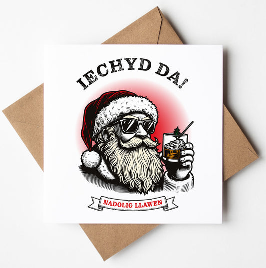 Illustration of Santa wearing sunglasses and holding a drink, with "Iechyd Da!" above and "Nadolig Llawen" below—perfect as a Welsh Christmas card. The backdrop is white with a brown envelope, ideal for worldwide shipping.