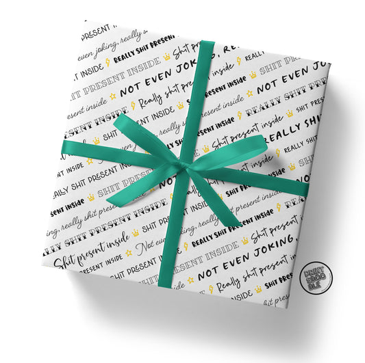 A gift box wrapped in the Shit Present Gift Wrap (Folded Sheet) displays playful and humorous text in black and gold, such as "SHIT PRESENT" and other whimsical phrases. It is adorned with a green ribbon and bow, along with a small circular sticker on the bottom right corner.