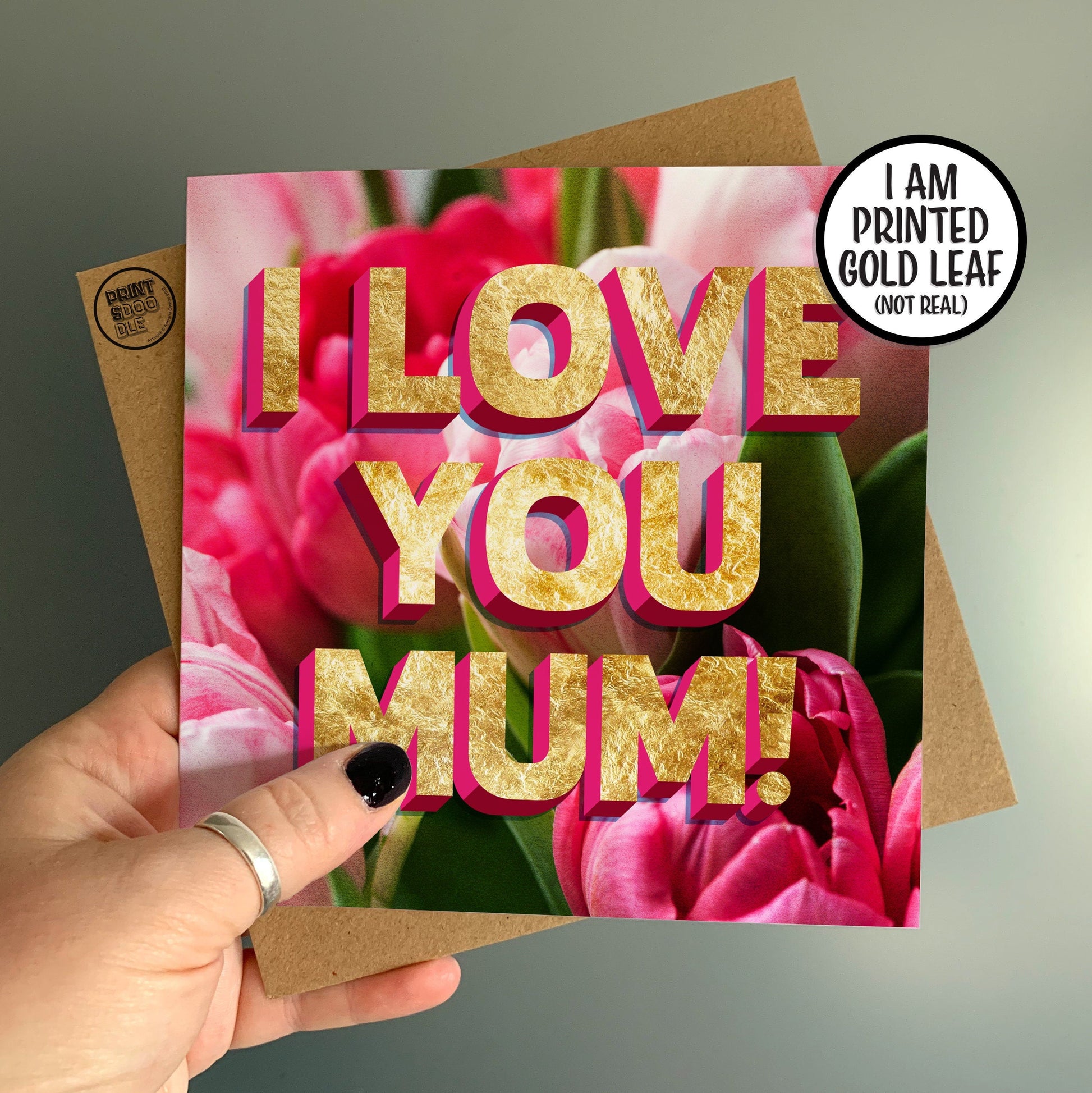 A hand holds the I Love You Mum Tulip Card, featuring vibrant pink and red tulips that make it an ideal floral Mother's Day card. The bold gold lettering adds warmth and love to the design. A sticker indicating "I AM PRINTED GOLD LEAF (NOT REAL)" is present, with a brown envelope seen in the background.